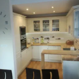 Jasna kuchnia w kształcie litery U z drewnianym blatem i barkiem, widok zza czarnych krzeseł z wysokim oparciem.