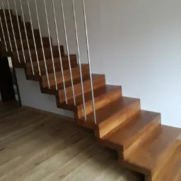 Drewniane schody z metalowymi barierkami, bez podstopnic, prowadzące w górę przy białej ścianie, oświetlone naturalnym światłem.