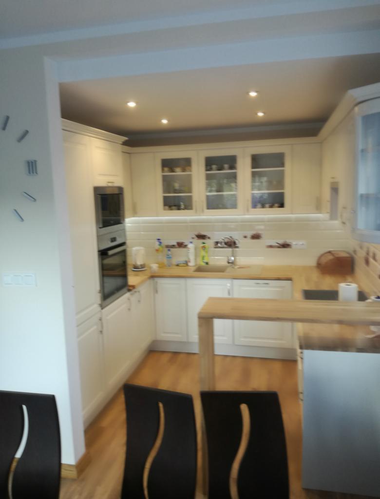 Jasna kuchnia w kształcie litery U z drewnianym blatem i barkiem, widok zza czarnych krzeseł z wysokim oparciem.