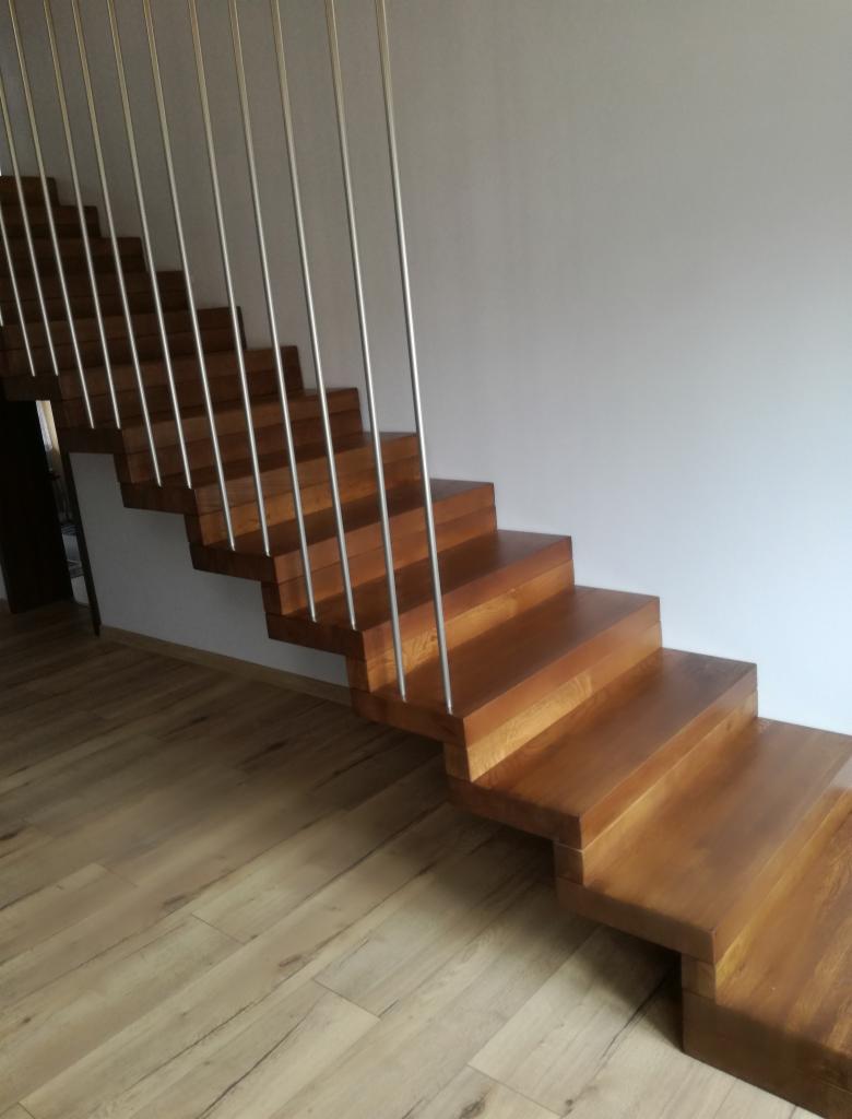 Drewniane schody z metalowymi barierkami, bez podstopnic, prowadzące w górę przy białej ścianie, oświetlone naturalnym światłem.