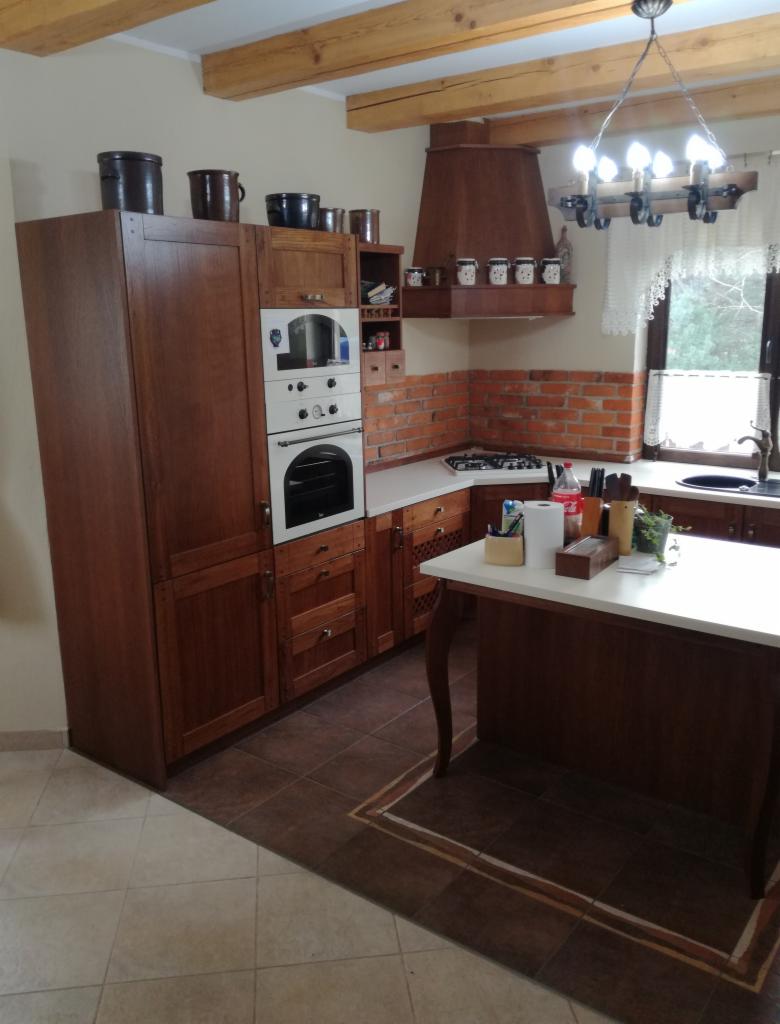 Rustykalna kuchnia z drewnianymi belkami stropowymi, ceglaną ścianą nad blatem, drewnianymi szafkami i wyspą z białym blatem.