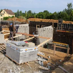 Budowa fundamentów domu jednorodzinnego: widoczne zbrojenie, szalunki, pracownicy na deskach, oraz palety z bloczkami PEBEK na terenie budowy.