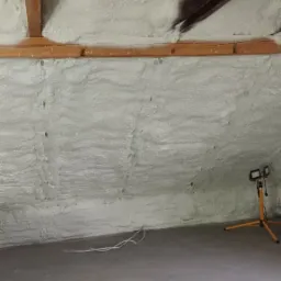 Poddasze po ociepleniu pianką poliuretanową, widoczne drewniane elementy konstrukcyjne oraz lampa budowlana na trójnogu.