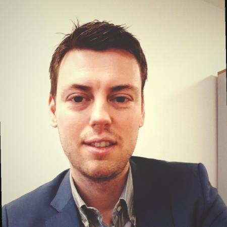 Młody mężczyzna w niebieskiej marynarce i koszuli w paski, uśmiecha się do kamery na jasnym tle. Portret biznesowy.