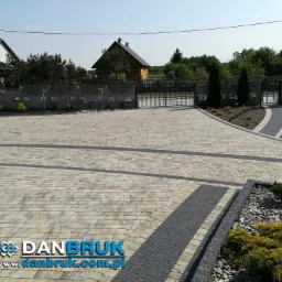 Szeroki podjazd z kostki brukowej w odcieniach beżu i szarości, z dekoracyjnymi pasami z drobnego kamienia, prowadzący do bramy wjazdowej i furtki, otoczony zadbaną zielenią i elementami małej...