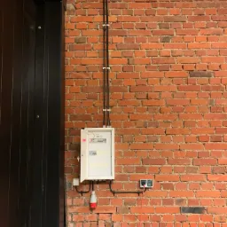 Skrzynka bezpiecznikowa z otwartymi drzwiczkami zamontowana na czerwonej ścianie z cegły, widoczne przewody w peszlach, gniazdko elektryczne i wtyczka z czerwonym kablem zasilającym.