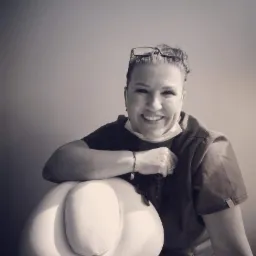 Uśmiechnięta kobieta w uniformie medycznym i okularach na głowie, oparta o biały fotel zabiegowy, zdjęcie czarno-białe.