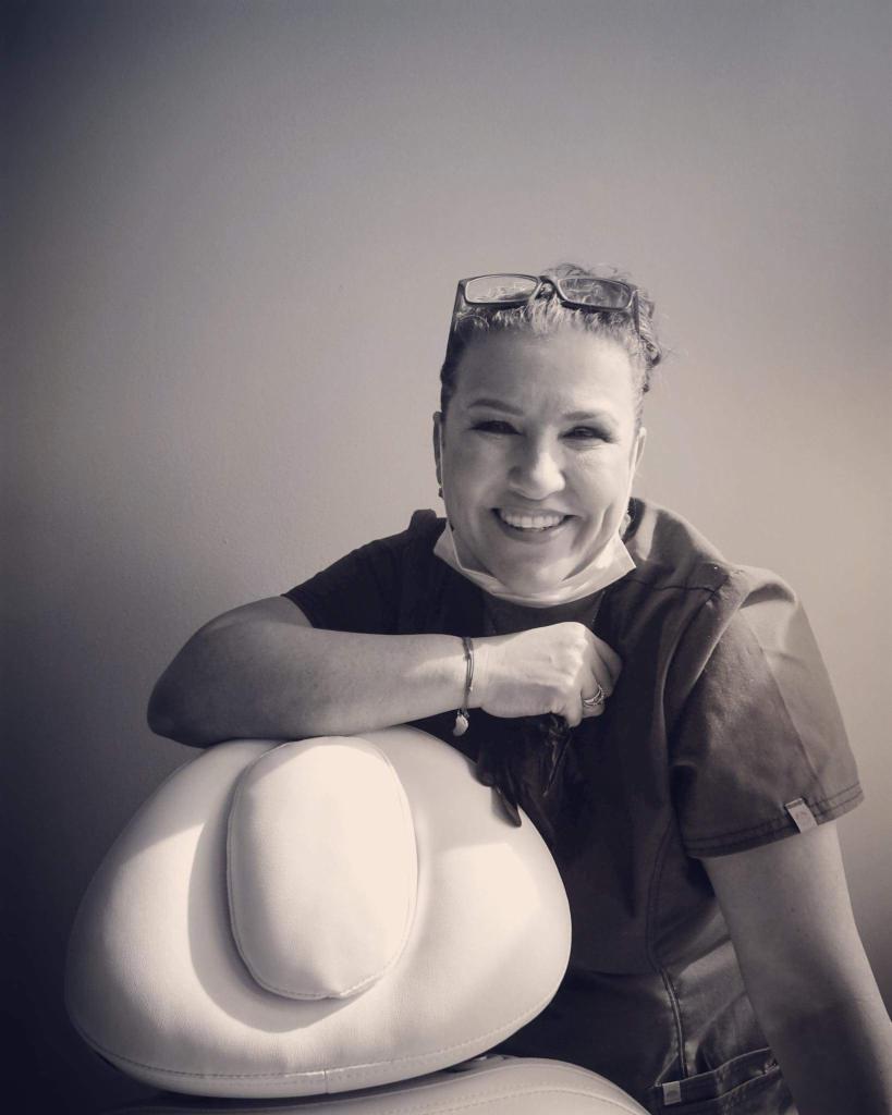 Uśmiechnięta kobieta w uniformie medycznym i okularach na głowie, oparta o biały fotel zabiegowy, zdjęcie czarno-białe.