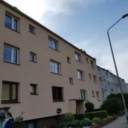 Wielopiętrowy budynek mieszkalny z elewacją w odcieniach beżu, widoczny z perspektywy ulicy, z elementami zieleni i latarnią uliczną. Elewacja po renowacji.