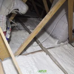 Pracownik w kombinezonie ochronnym aplikuje białą pianę izolacyjną między krokwiami poddasza, widoczne drewniane elementy konstrukcji dachu i fragmenty instalacji elektrycznej.