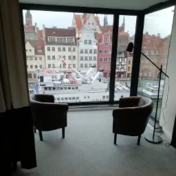 Wnętrze pokoju w Gdańsku z widokiem na Motławę i zabytkową zabudowę przez duże okno, z dwoma fotelami i lampą podłogową, zasłonięte częściowo zasłonami.