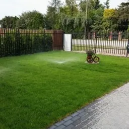 Automatyczny system nawadniania trawnika w ogrodzie, zraszacz w akcji, ozdobny kwietnik w kształcie roweru, ogrodzenie panelowe.