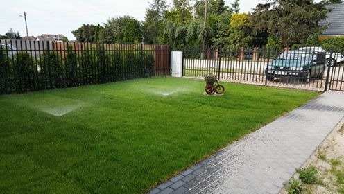 Automatyczny system nawadniania trawnika w ogrodzie, zraszacz w akcji, ozdobny kwietnik w kształcie roweru, ogrodzenie panelowe.