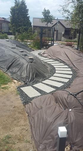 Ścieżka z prostokątnych płyt ułożona na czarnym żwirze, prowadząca przez teren ogrodu zabezpieczony czarną i brązową agrowłókniną, w tle zabudowa jednorodzinna.