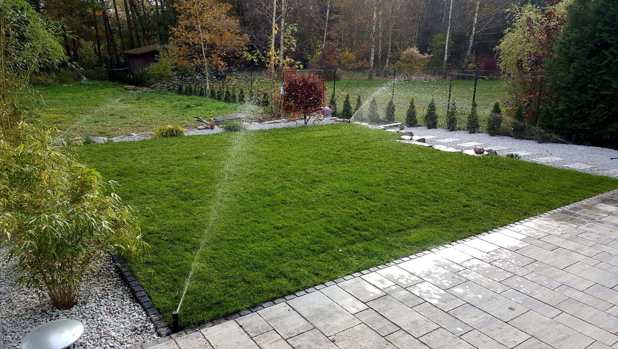 Automatyczny system nawadniania trawnika w ogrodzie, zraszacze w trakcie pracy, widok na równo przystrzyżoną trawę, kamienne ścieżki i obrzeża, oraz kostkę brukową na tarasie.