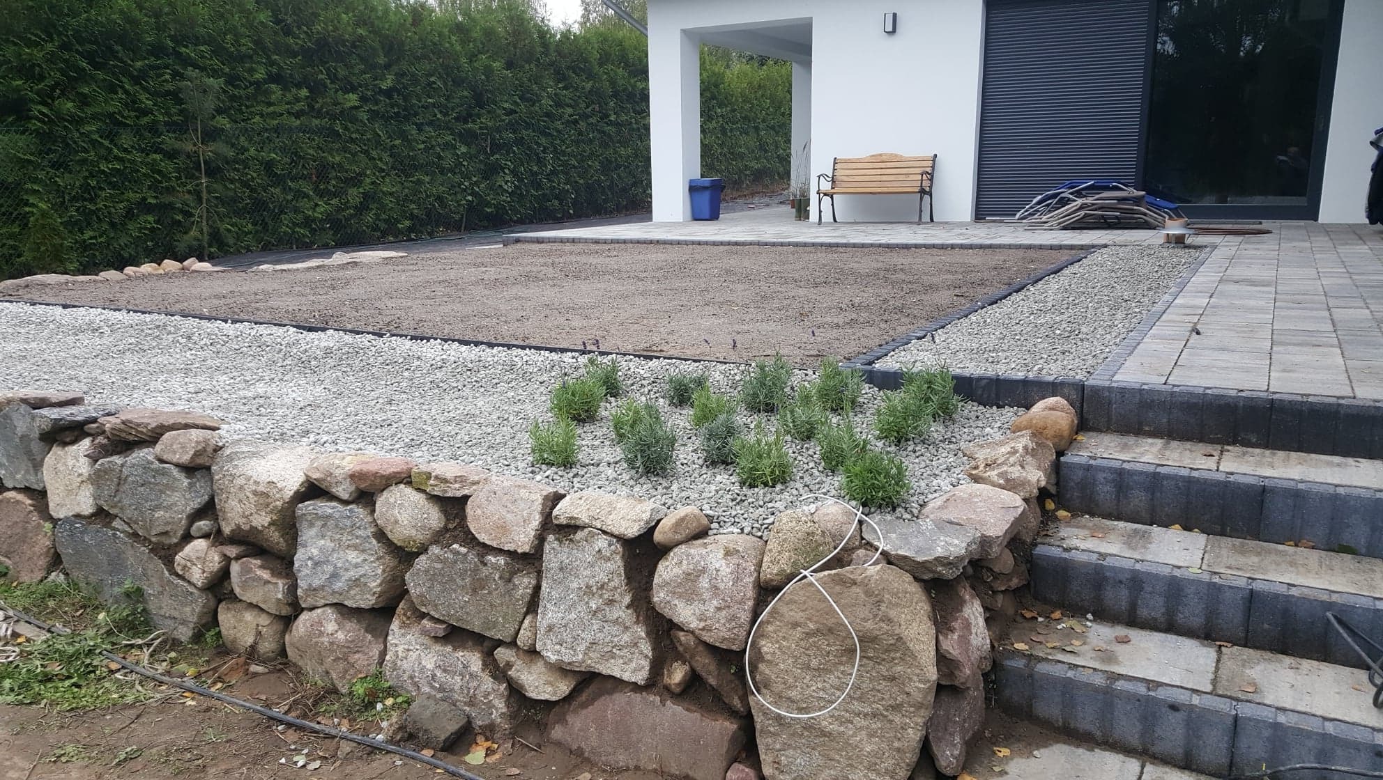 Etap zakładania ogrodu: kamienny murek oporowy, żwirowe obrzeże, przygotowany teren pod trawnik i nasadzenia lawendy, kostka brukowa na tarasie i schodach.
