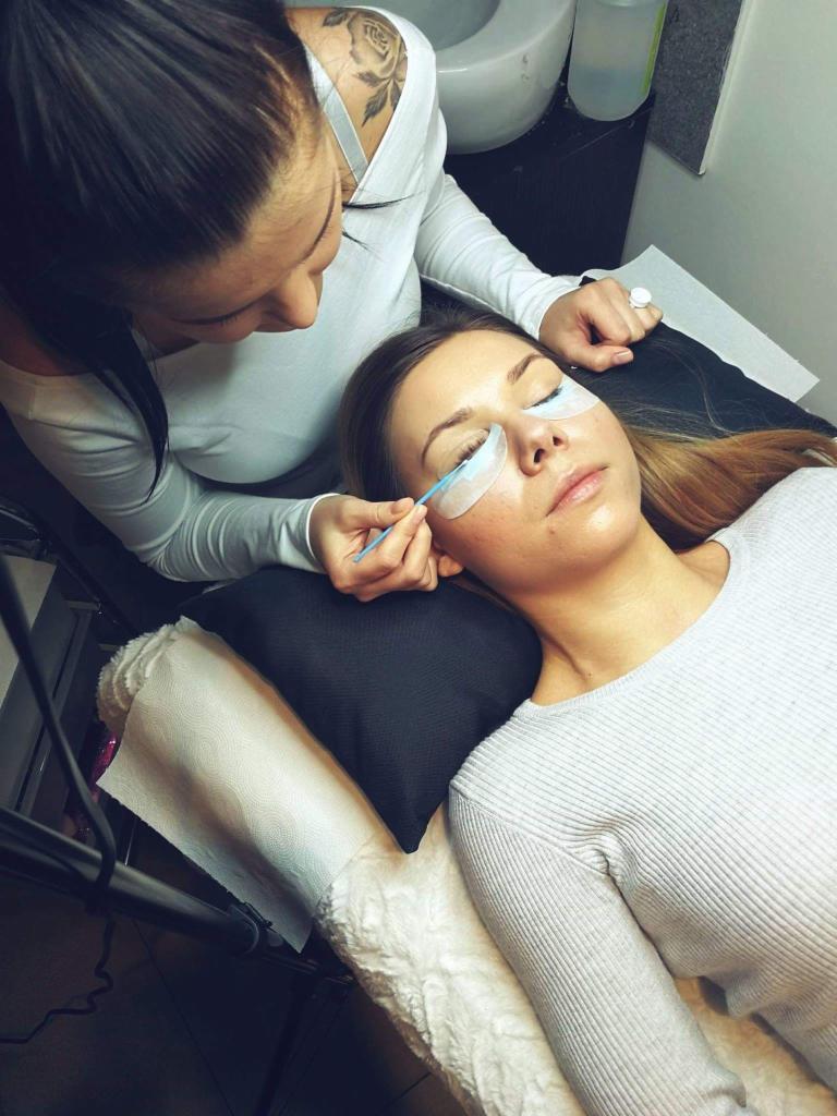 Kobieta wykonuje zabieg na rzęsach klientki leżącej na leżance, z plasterkami ochronnymi pod oczami, widoczne narzędzie do aplikacji, w tle umywalka i kosmetyki.