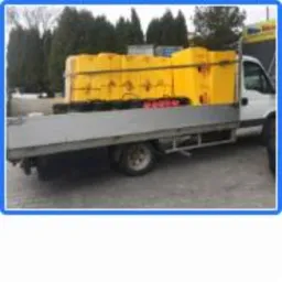 Samochód dostawczy z żółtymi paczkami materiału izolacyjnego na pace, widoczne drzewa w tle.