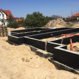 Wykopane i zabezpieczone fundamenty domu jednorodzinnego z widocznymi elementami szalunku, rurami kanalizacyjnymi i paletami w tle.