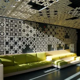 Wnętrze z dekoracyjnymi panelami ażurowymi na ścianie i suficie, zielona sofa modułowa, roślina w donicy, gra świateł na podłodze.