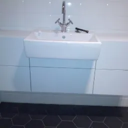 Biała, prostokątna umywalka ceramiczna z chromowaną baterią, zamontowana na szafce z białymi, gładkimi frontami, na tle białych płytek ściennych i heksagonalnej, ciemnej podłogi. Na umywalce leży...