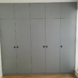 Szara, zabudowana szafa z ciemnymi, minimalistycznymi uchwytami, dopasowana do wysokości pomieszczenia, z widocznym fragmentem drewnianej podłogi i oknem z białymi ramami.