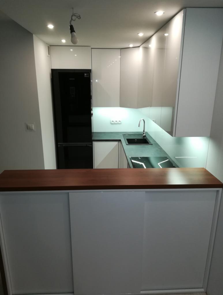 Nowoczesna, minimalistyczna kuchnia z białymi, błyszczącymi frontami, czarnym blatem i drewnianym barkiem. Widoczna czarna lodówka i oświetlenie podszafkowe.