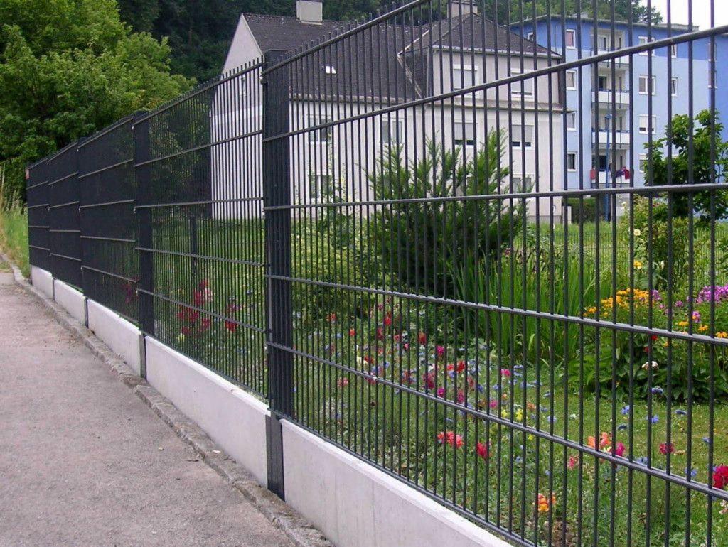 Ciemne, stalowe ogrodzenie z paneli siatkowych na betonowej podmurówce, oddzielające chodnik od rabaty kwiatowej przed budynkiem mieszkalnym.