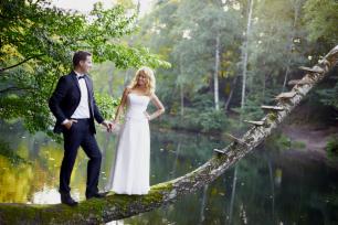 Para młoda w strojach ślubnych stoi na powalonym drzewie nad wodą, w tle zielony las.