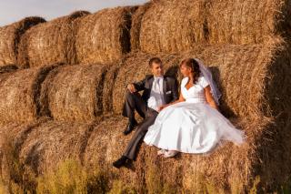 Para młoda w strojach ślubnych siedzi na stogu siana w Sosnowcu, ujęcie z dołu, widoczne niebo w tle.