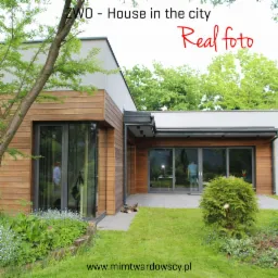 Nowoczesny budynek usługowy z drewnianą elewacją i dużymi przeszkleniami, otoczony zielonym ogrodem z krzewami i drzewami; na tarasie widać leżącego kota.