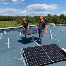 Las24.pl lasogród Konrad Namysło Filip Namysło sp.j. - Dwóch monterów w pomarańczowych kaskach instaluje panele fotowoltaiczne na płaskim dachu w słoneczny dzień, widoczne metalowe konstrukcje montażowe i las w tle.