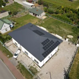 Widok z drona na dom z ciemnoszarym dachem i panelami fotowoltaicznymi, biała elewacja, teren wokół wysypany jasnym żwirem, biały samochód dostawczy zaparkowany obok, w tle inne domy i zieleń.