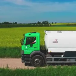 Zielona cysterna z logo Naftpol na tle kwitnącego pola rzepaku, widok z boku na polnej drodze.