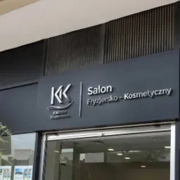 Elegancki szyld salonu fryzjersko-kosmetycznego z logo 'KK' i nazwą 'Katarzyna Krzyżanowska' na ciemnoszarym tle budynku.