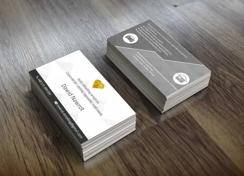 Dwa stosy wizytówek na drewnianym blacie, jedna z białym tłem i złotym logo firmy auto detailingu, druga z szarym tłem i ikonami usług.