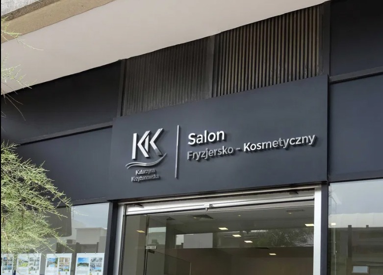 Elegancki szyld salonu fryzjersko-kosmetycznego z logo 'KK' i nazwą 'Katarzyna Krzyżanowska' na ciemnoszarym tle budynku.