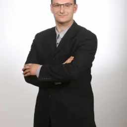 Mężczyzna w okularach i czarnym garniturze, z założonymi rękami na piersi, pozuje na jasnym tle.