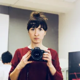 Kobieta z upiętymi włosami trzyma aparat Canon, patrząc w obiektyw. Widoczne odbicia światła w soczewce. Ubrana w bordową bluzkę, z czerwonym manicure. Tło stanowią białe i szare ściany.