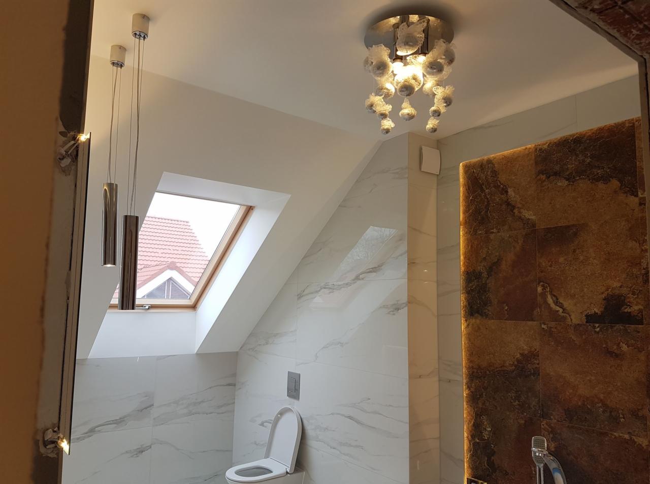 Ukończona łazienka na poddaszu z oknem dachowym, marmurowymi płytkami ściennymi, wiszącą toaletą i dekoracyjną lampą sufitową z elementami szkła