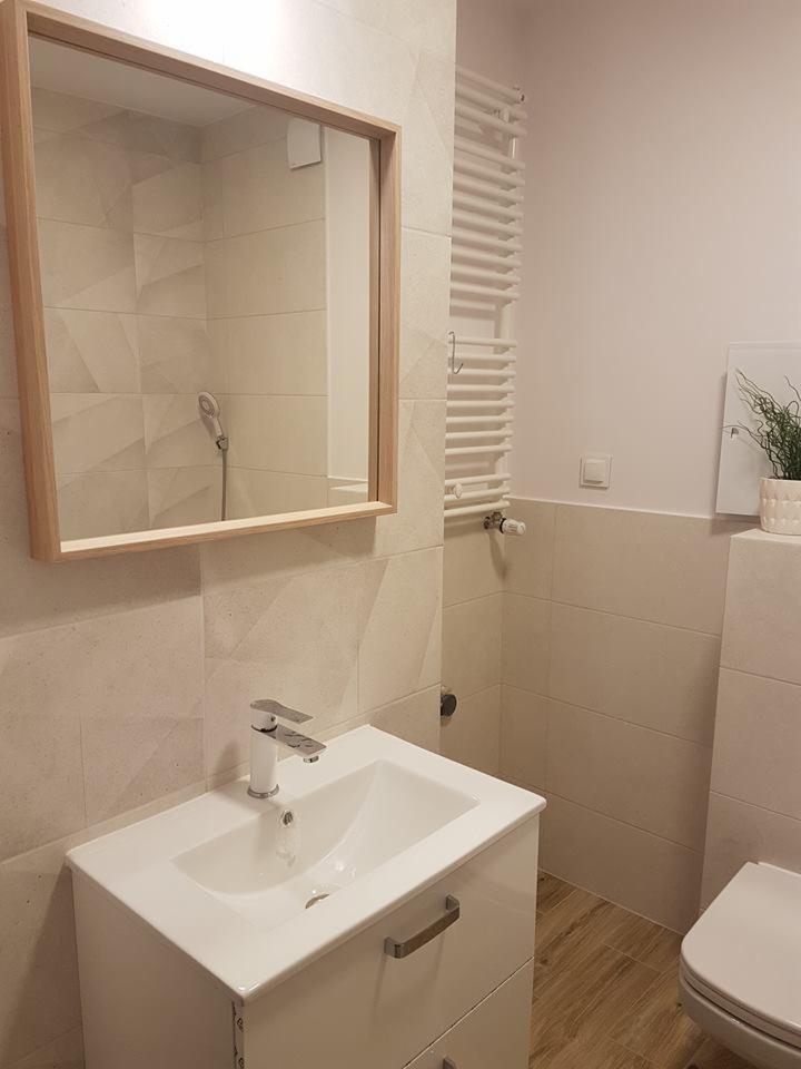 Jasna łazienka z beżowymi płytkami o geometrycznym wzorze, drewnianą ramą lustra i białymi meblami łazienkowymi, grzejnikiem drabinkowym i akcentem roślinnym.