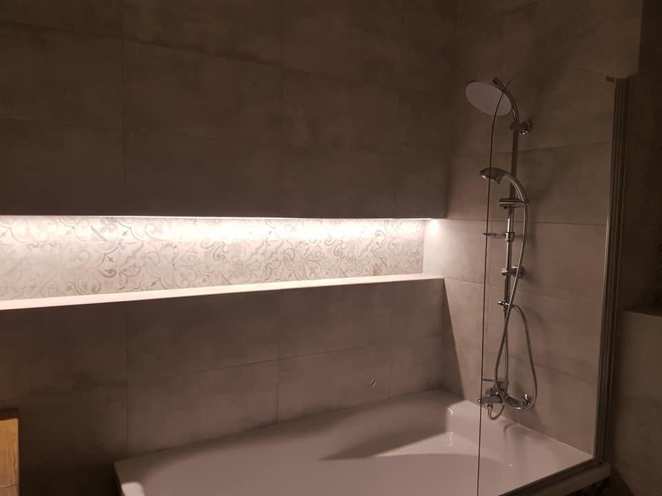 Nowoczesna łazienka z wanną, szklanym parawanem i chromowaną baterią prysznicową. W ścianie wnęka z podświetlaną dekoracyjną płytką.