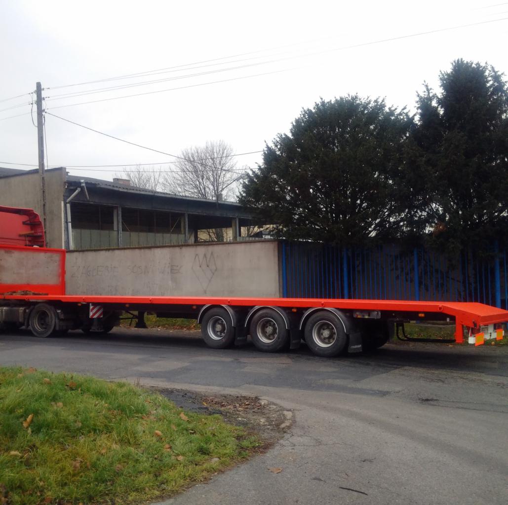 Czerwona, pusta naczepa niskopodwoziowa z graffiti 'Zaglebie Sosnowiec' na bocznej ścianie, zaparkowana na asfaltowej drodze, z budynkiem przemysłowym w tle.