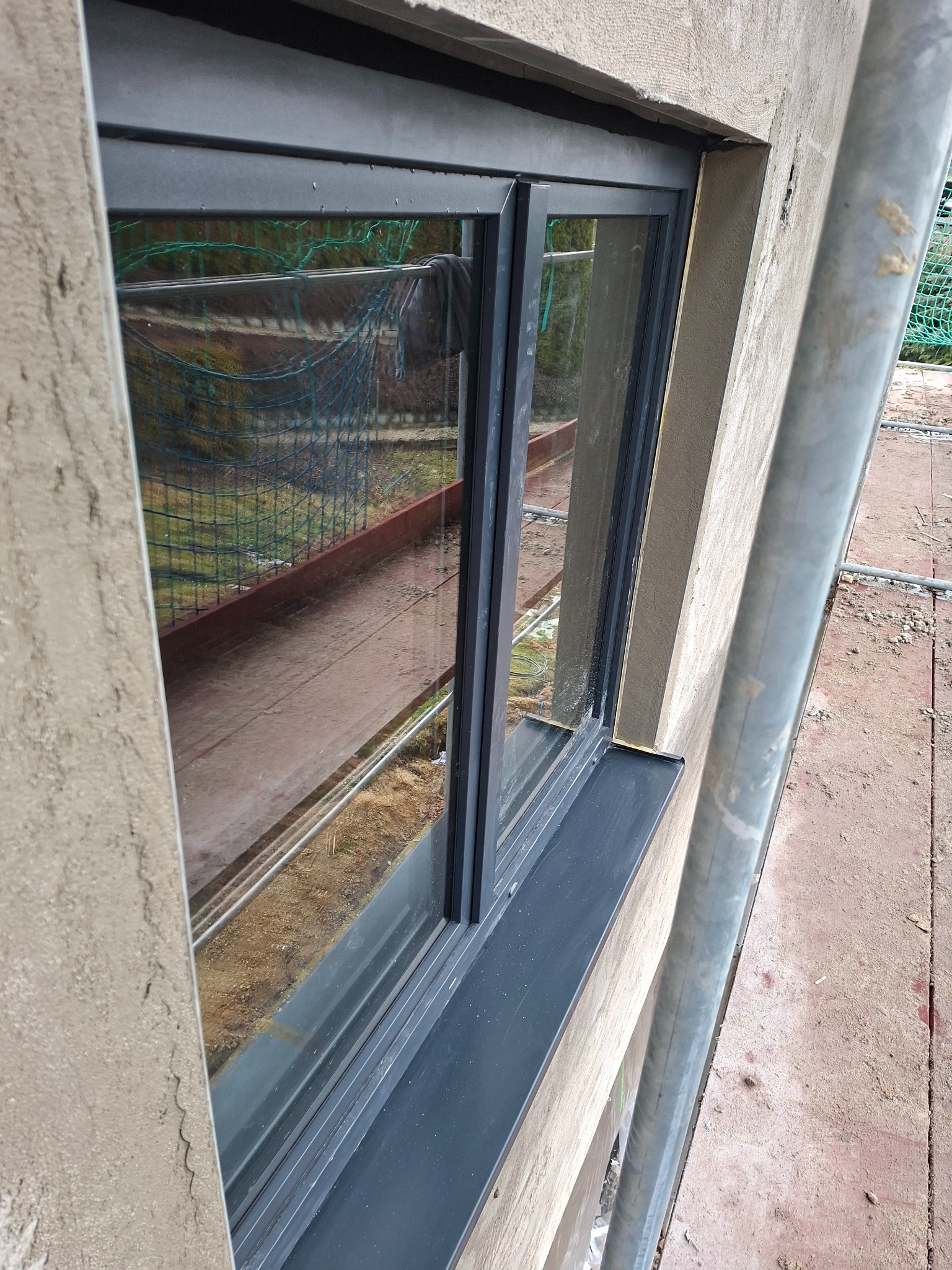 Montaż okna z czarną ramą w surowym murze. Widoczny fragment rusztowania. Ujęcie z bliska, podkreślające detale wykończenia i surowość otoczenia.