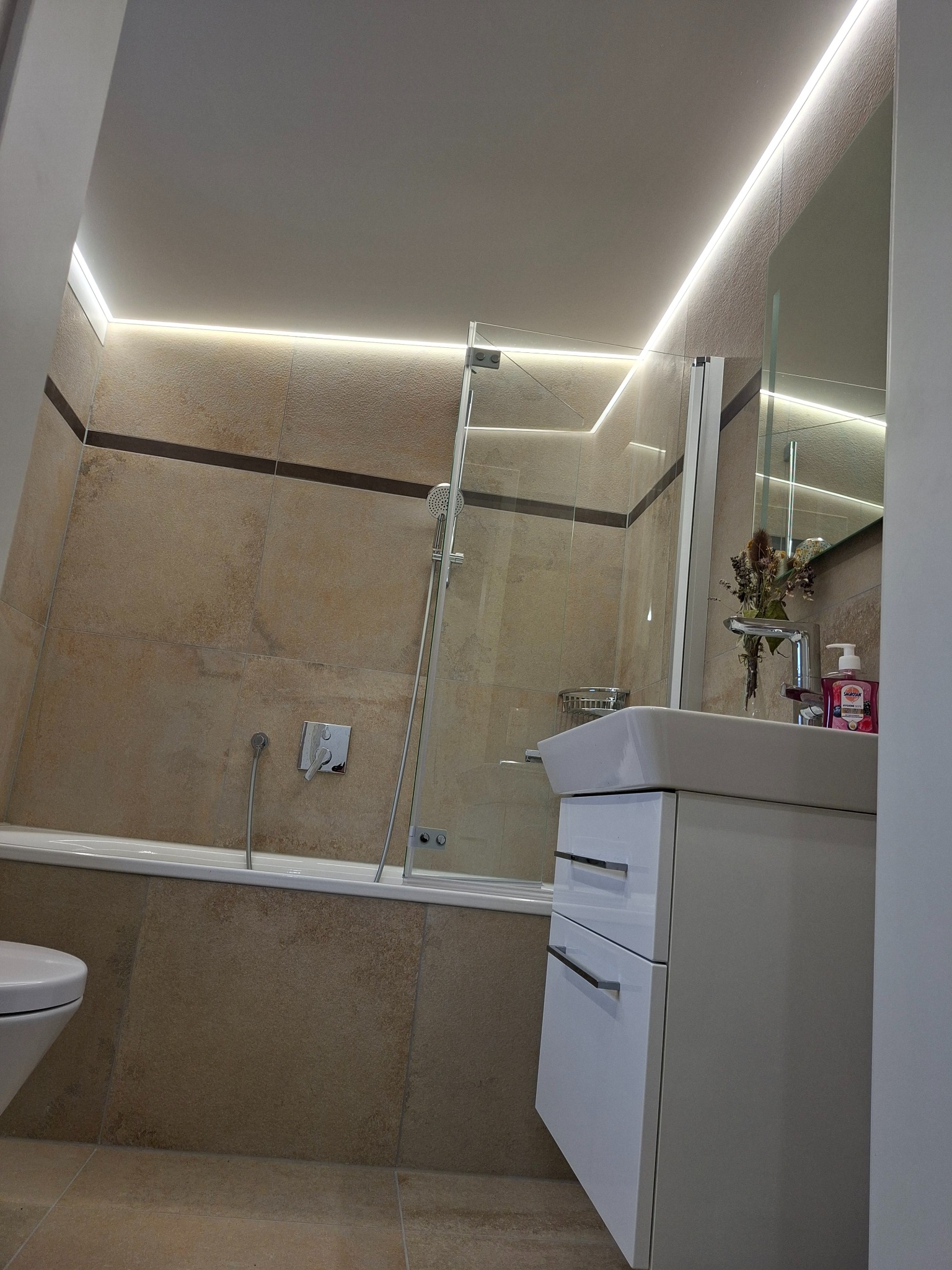 Wykończona łazienka z beżowymi płytkami, podświetleniem LED, białą szafką pod umywalką i szklanym parawanem nawannowym. Nowoczesny design.