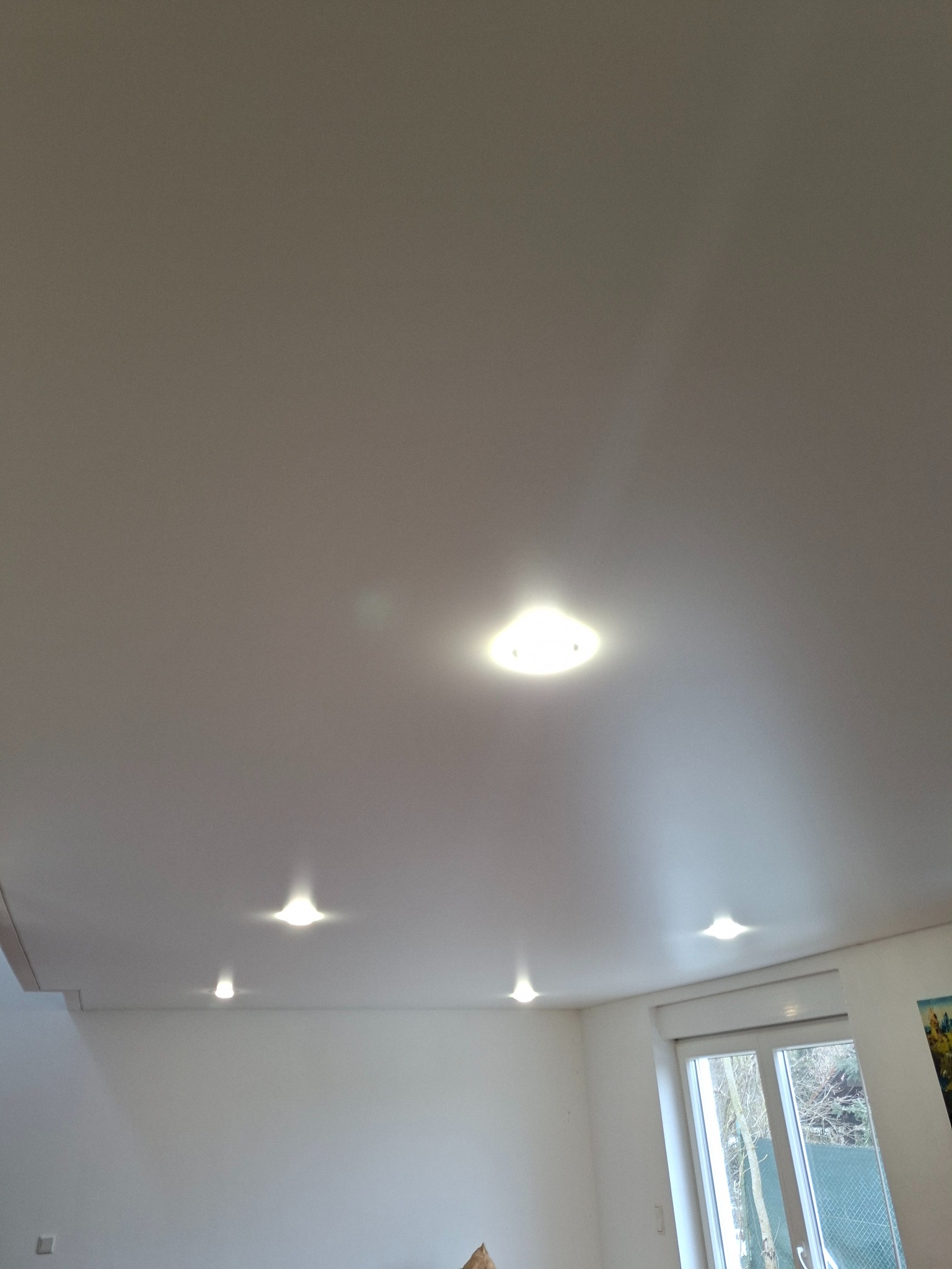 Biały sufit podwieszany z wbudowanymi, punktowymi lampami LED, widok z dołu, fragment okna z roletą w tle. Nowoczesne oświetlenie w jasnym wnętrzu.