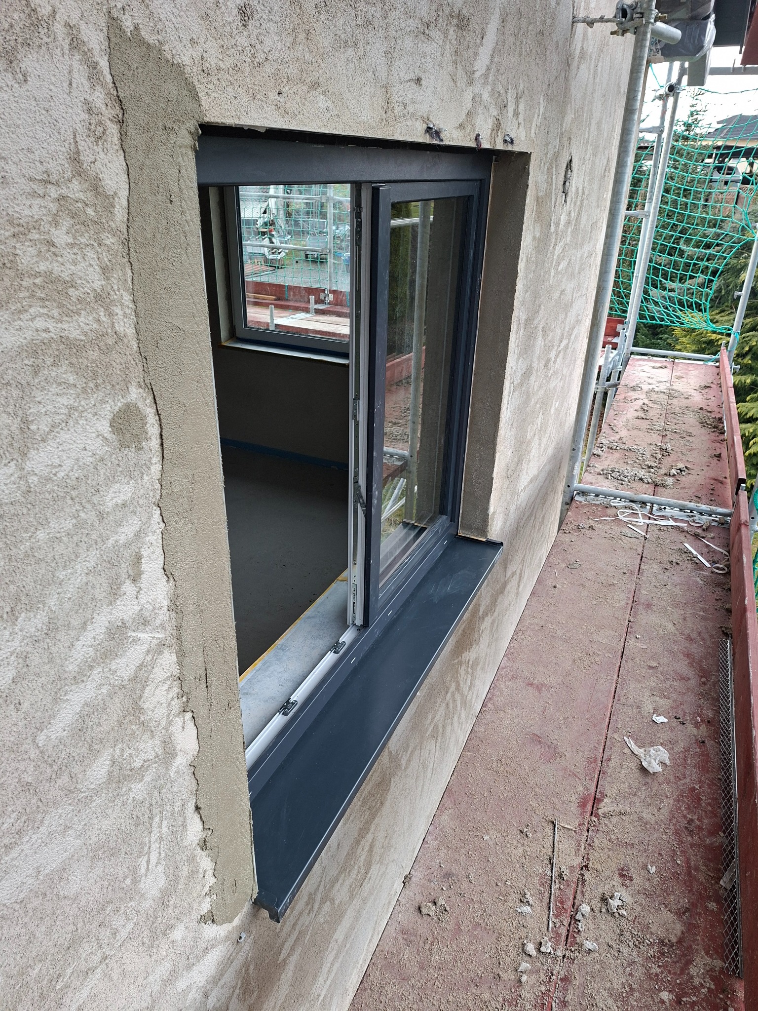 Montaż okna z szarą ramą w surowej elewacji. Widoczne rusztowanie i narzędzia. Etap prac wykończeniowych.