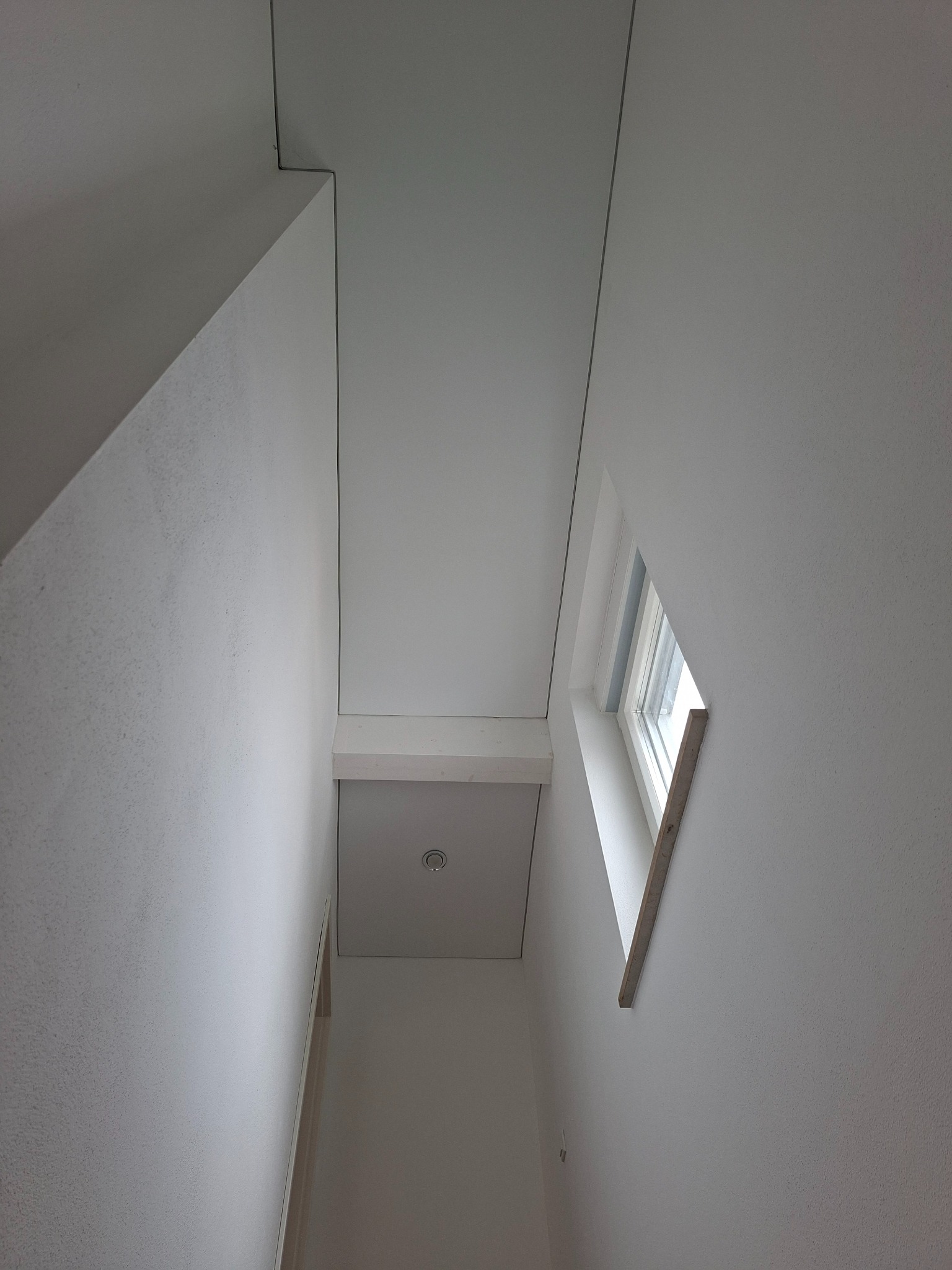 Wnętrze klatki schodowej z oknem o nietypowym kształcie, wnęka oświetlona naturalnym światłem. Ściany i sufit w odcieniach bieli, minimalistyczny design.