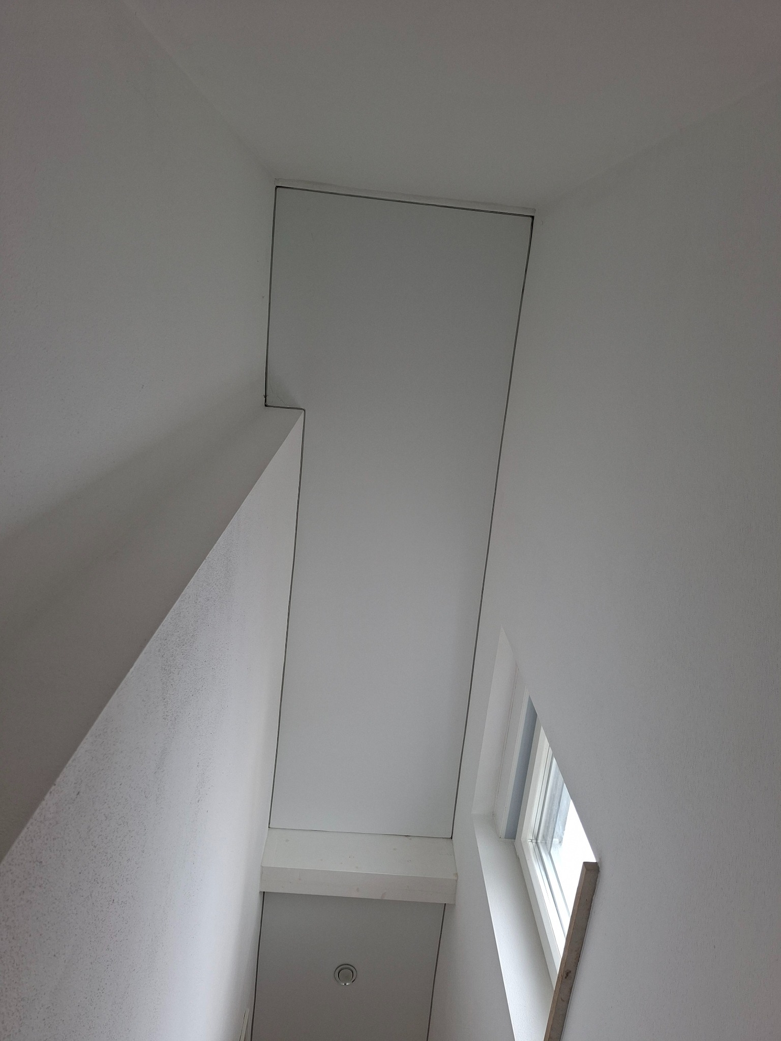 Białe wnętrze ze skośnym sufitem i oknem w kształcie trapezu. Widoczne połączenie ściany ze sufitem i fragment schodów. Minimalistyczny design.