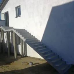 Surowe betonowe schody zewnętrzne prowadzące do wejścia w nowo otynkowanym budynku, z widocznym wiadrem na ziemi i częściowo odsłoniętą konstrukcją wsporczą.